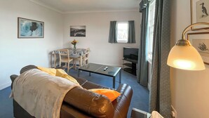 A living room with a brown leather sofa, a glass coffee table, a TV, a dining table with chairs, and a chandelier.