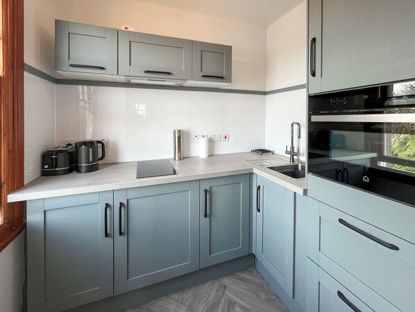 A modern kitchen with grey cabinets, a white countertop, a built-in oven, and a kettle.