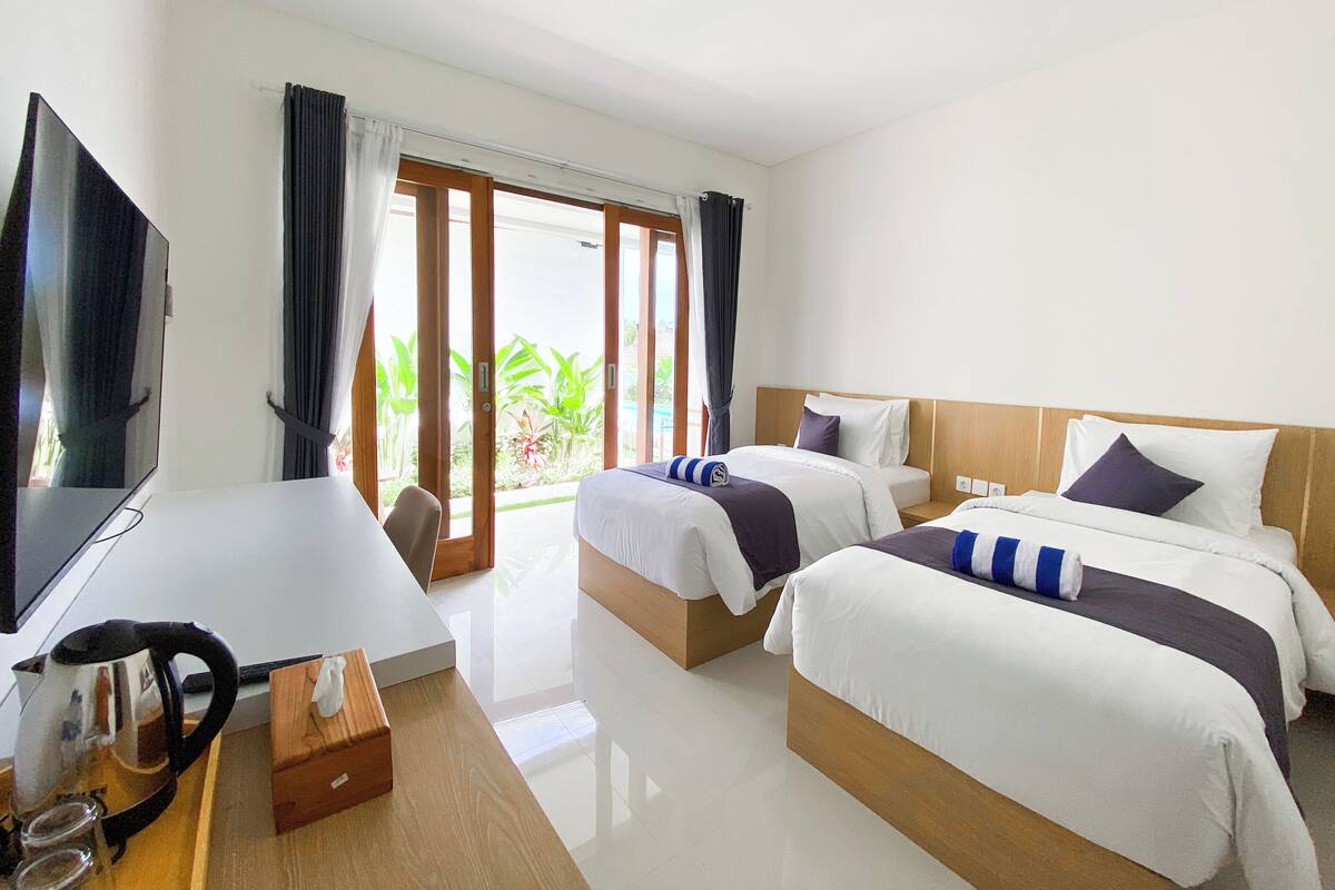 A hotel room with two beds, a TV, a desk, and a view of greenery outside.