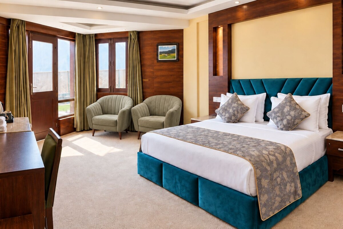 Habitación de hotel con una cama grande, dos sillones, un escritorio y un ventanal con cortinas.