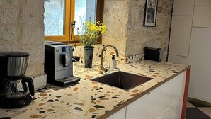 A kitchen with a stone wall, wooden beams, a window, a sink, a coffee maker, and a potted plant. - La Maison Soleil du Levant (Sérignac)