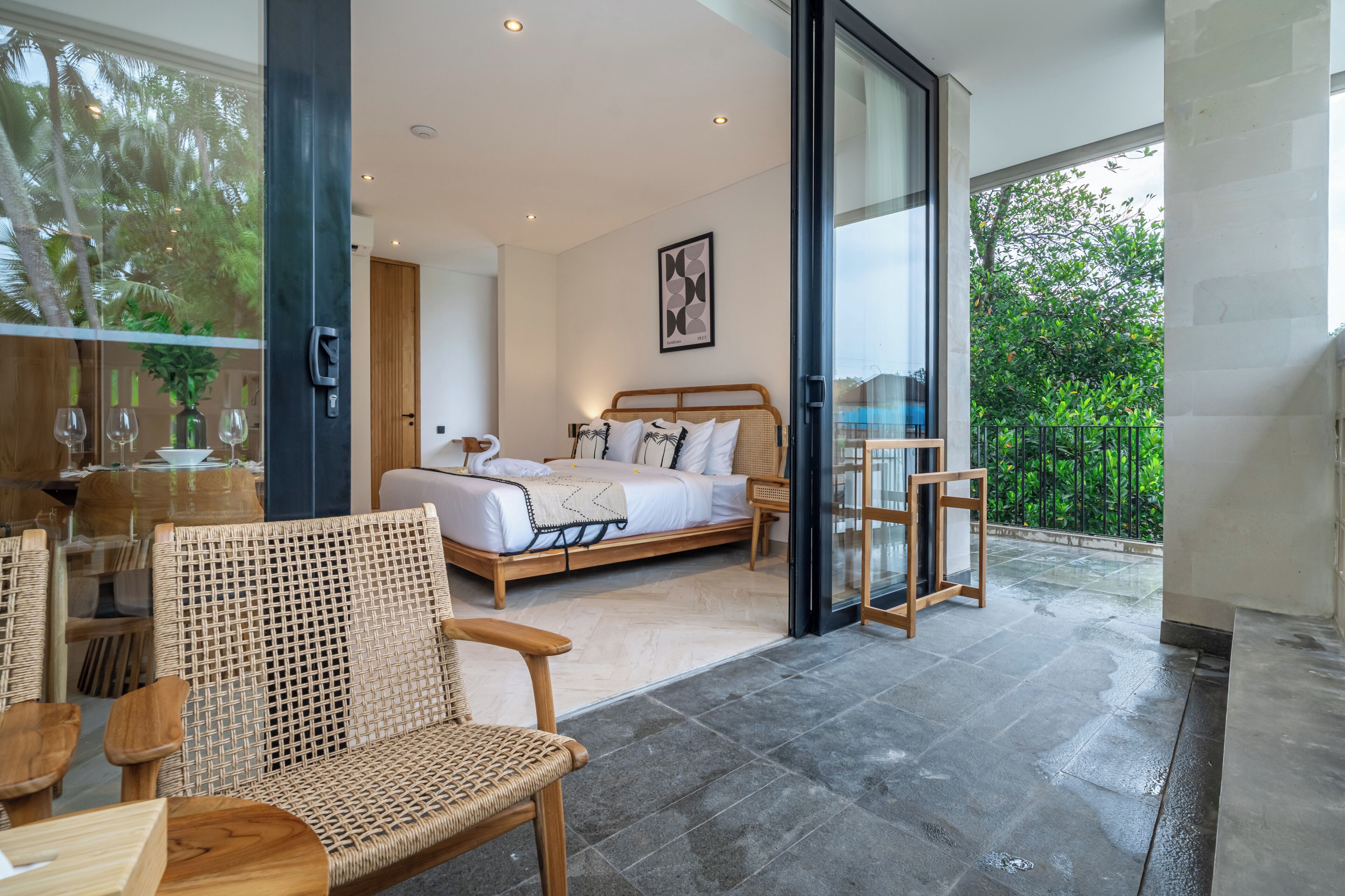 Une chambre moderne avec un lit, une porte vitrée donnant sur un balcon, des meubles en osier et un tableau encadré au mur.