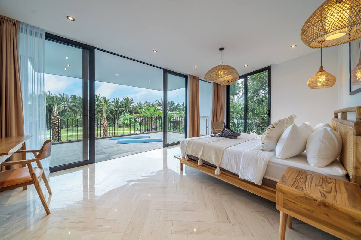 A modern bedroom with a large bed, a wooden bench, a desk, and a view of a lush garden through large windows.