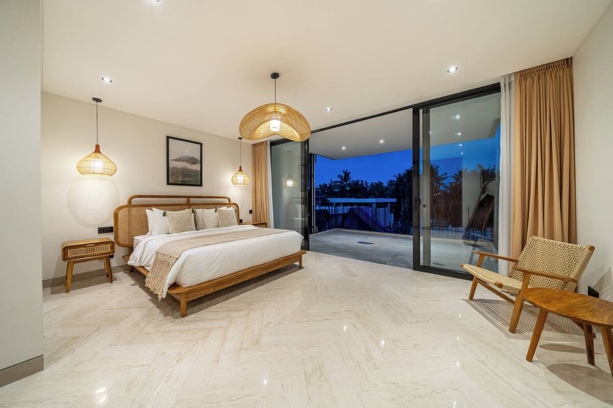 A modern bedroom with a large bed, bedside tables, a chair, and a glass door leading to a balcony.