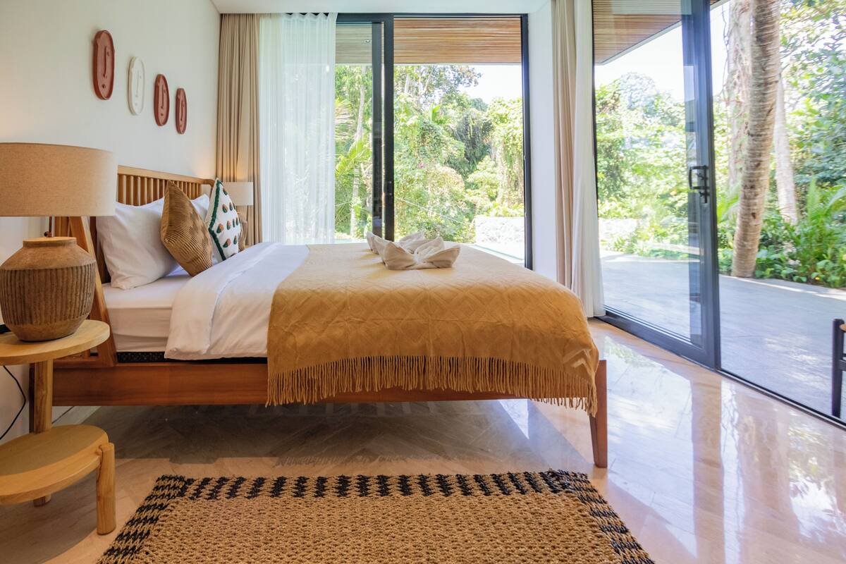 A modern bedroom with a large bed, a wooden headboard, a bedside table with a lamp, and a view of lush greenery through large windows.