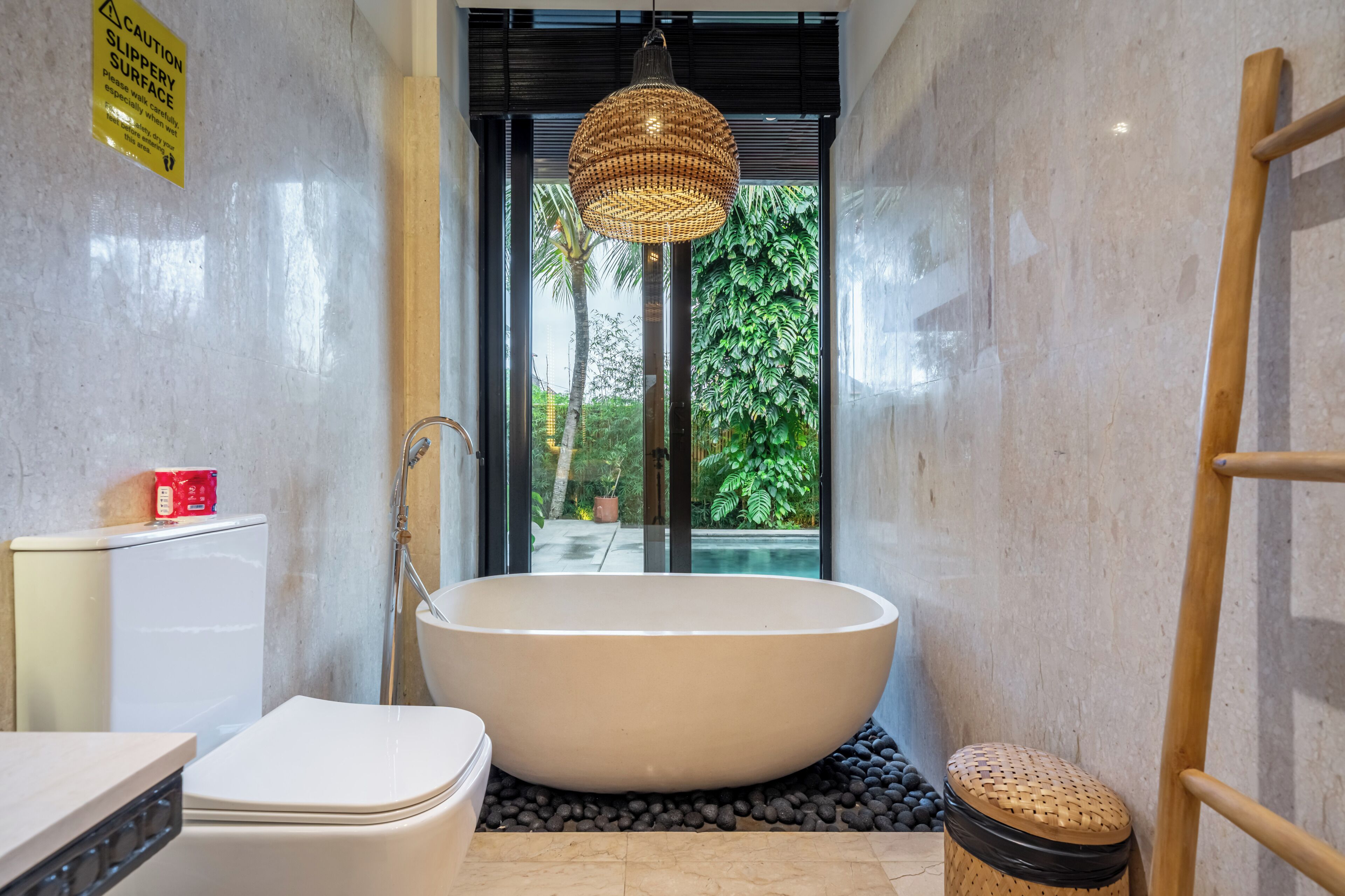 A bathroom with a large bathtub, a toilet, a sink, a red box on the toilet, and a window with a view of greenery.