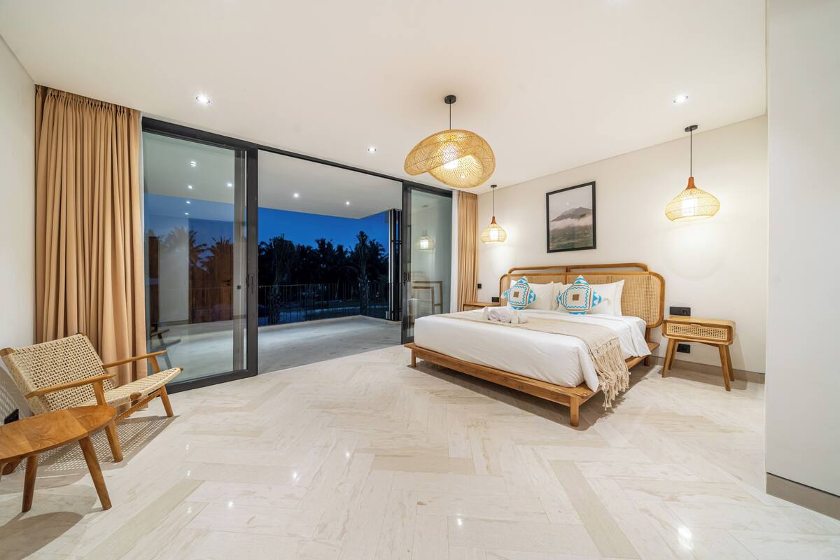 A bedroom with a large bed, a chair, a nightstand, and a view of the outdoors.