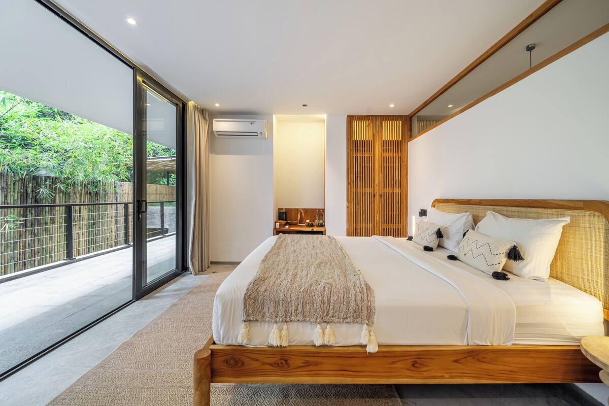 A modern bedroom with a large bed, a wooden headboard, a view of greenery outside, and a sliding glass door.