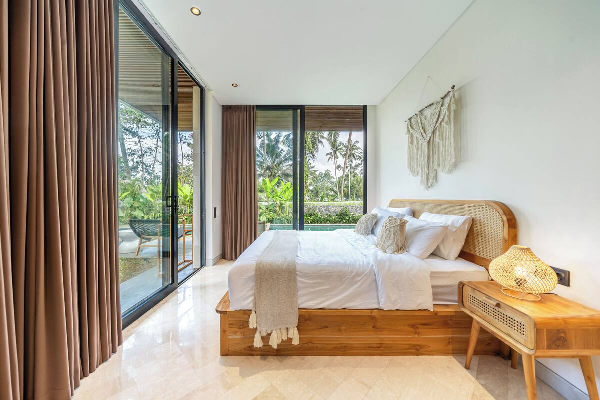 A modern bedroom with a large bed, a wooden nightstand, and a view of greenery through the window.