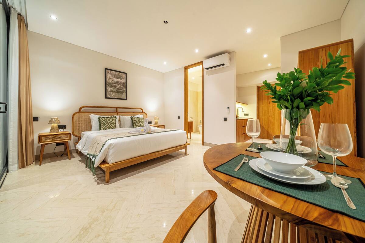 A hotel room with a bed, bedside table, lamp, and a dining area with a table set for two.