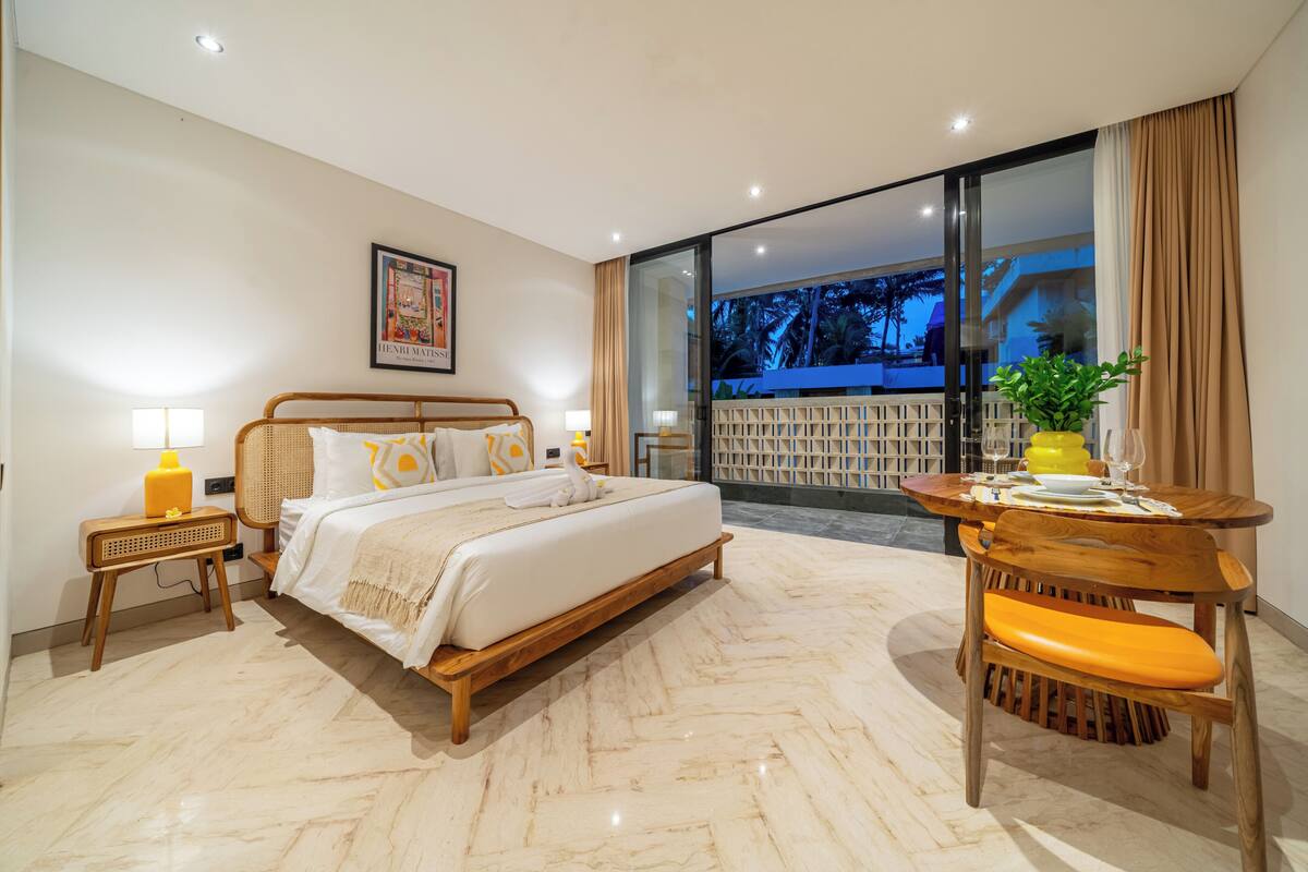 A bedroom with a bed, bedside table, lamp, and a view of the outside.