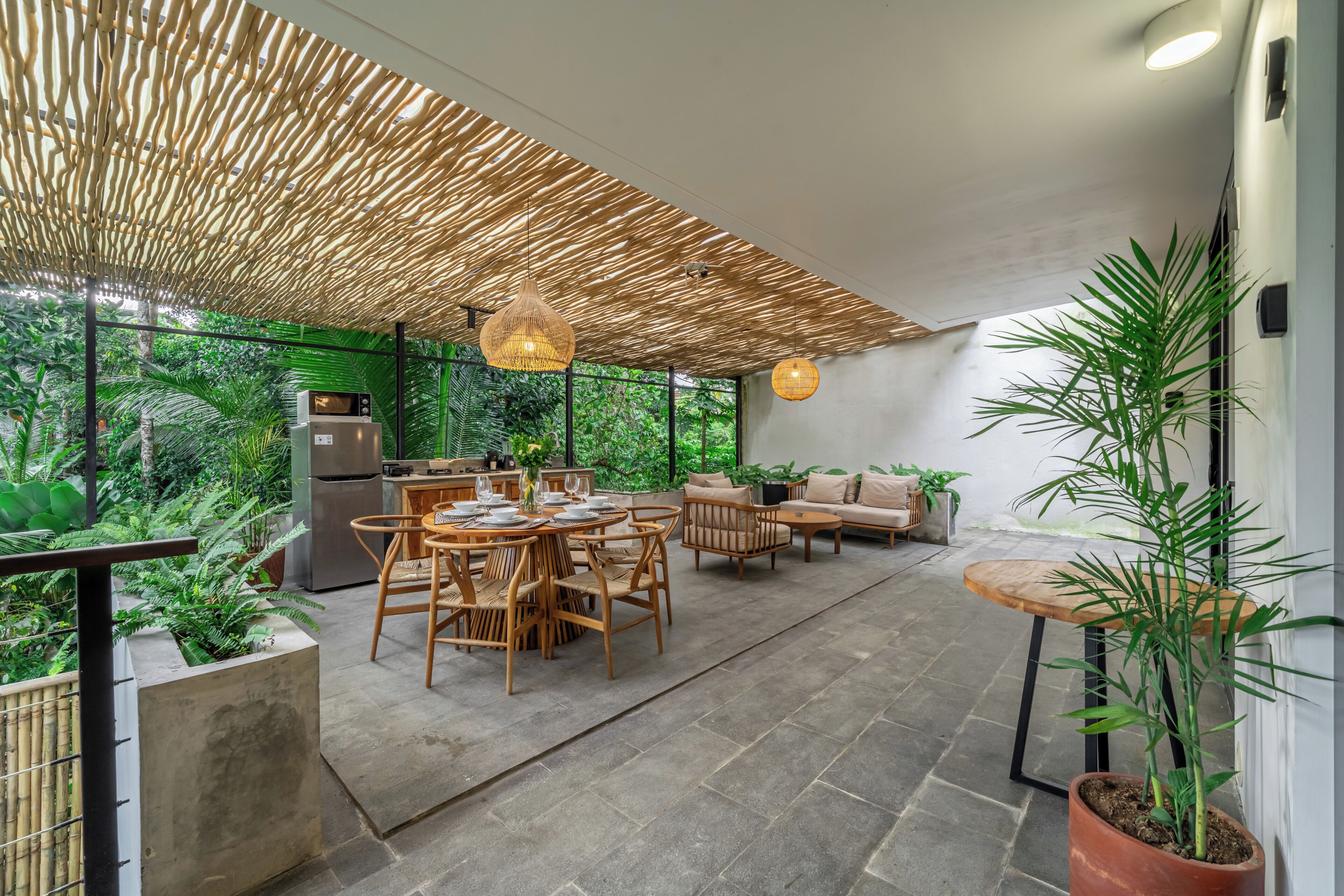 Un espacio exterior moderno con comedor, techo de bambú, cocina y zona de estar con sofá y mesa.