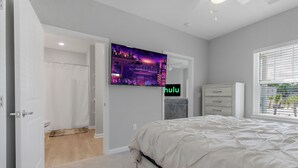 Ein Schlafzimmer mit einem Bett, einem an der Wand befestigten Fernseher, einem Fenster mit Jalousien und einer Tür zu einem anderen Raum.