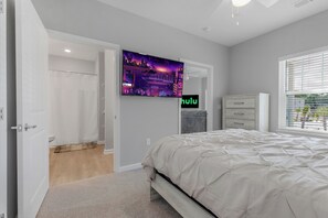 A bedroom with a bed, a TV mounted on the wall, a window with blinds, and a door leading to another room. - The Saraland (Mobile)