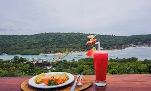 A table with a plate of food and a drink, overlooking a coastal landscape with greenery and a bridge.