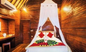 A wooden room with a bed under a canopy, a small table with a kettle, and a door.