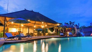 Un área junto a la piscina con sillones de descanso y sombrillas, una zona de estar cubierta con iluminación y una vista despejada de la piscina.