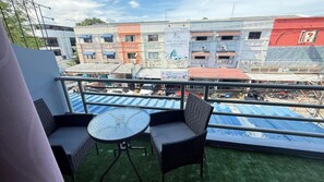 A balcony with a round table and two chairs, overlooking a cityscape with buildings and a 7-Eleven store. - Chalong Paradise Guesthouse (Rawai)