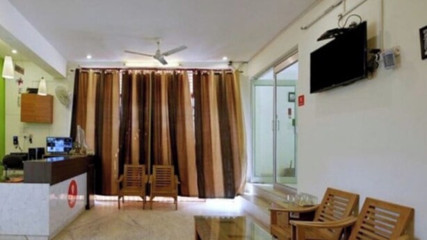 A hotel lobby with a reception desk, wooden chairs, a glass-top table, curtains, a ceiling fan, and a TV mounted on the wall. - Hotel GSA and BANQUET (Haridwar)