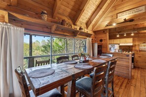 A wooden cabin interior with a dining table, chairs, a kitchen area, and a large window overlooking greenery. - 2 Cabins + 2 Hot Tubs + Fire Pit | Smokies Views | Elianah's & Maggie's Cabin (Townsend)