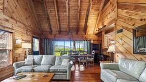 A wooden cabin interior with a sofa, coffee table, dining table, chairs, and a view of the outdoors. - 6BR Cabin Trio + Hot Tubs | Quiet Side of Smokies | Isaiah's, Jack's & Elijah's (Townsend)