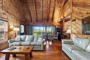 A wooden cabin interior with a sofa, coffee table, dining table, chairs, and a view of the outdoors.
