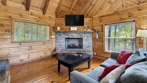 A log cabin interior with a stone fireplace, wooden walls, a flat-screen TV, and a sofa. - 6BR Cabin Trio + Hot Tubs | Quiet Side of Smokies | Isaiah's, Jack's & Elijah's (Townsend)