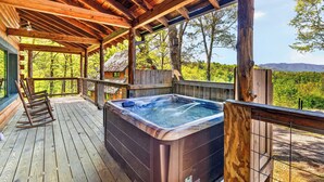 A wooden deck with a hot tub, rocking chairs, and a view of a forested area. - 6BR Cabin Trio + Hot Tubs | Quiet Side of Smokies | Isaiah's, Jack's & Elijah's (Townsend)