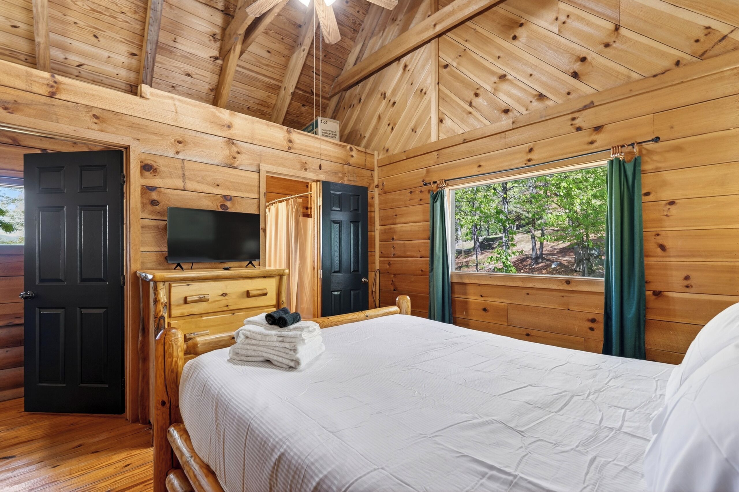 A wooden cabin room with a bed, a TV, a dresser, and a window with curtains.