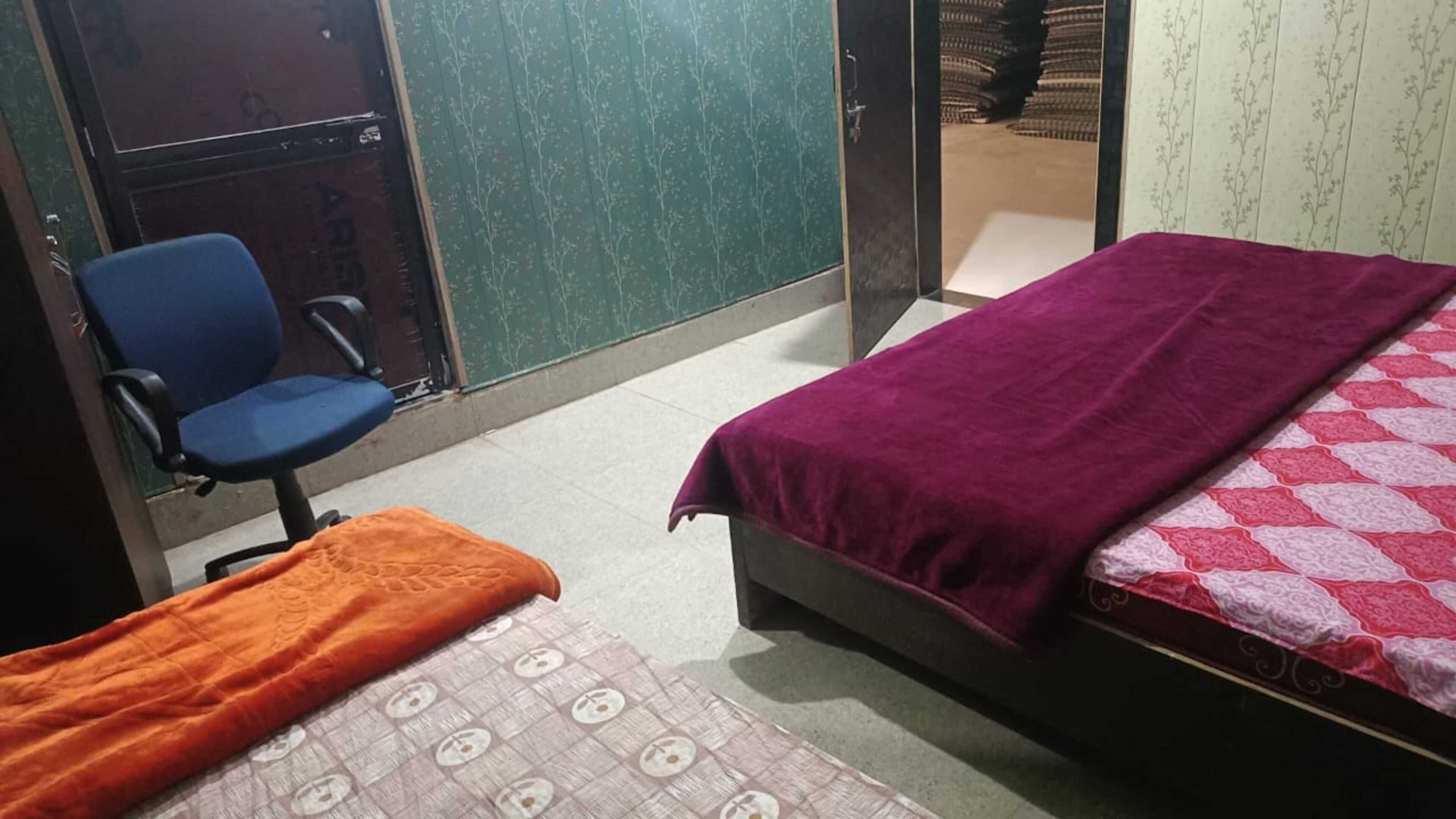 A room with two beds, one with a purple blanket and another with an orange blanket. There is a blue chair and a bed with a patterned bedspread.