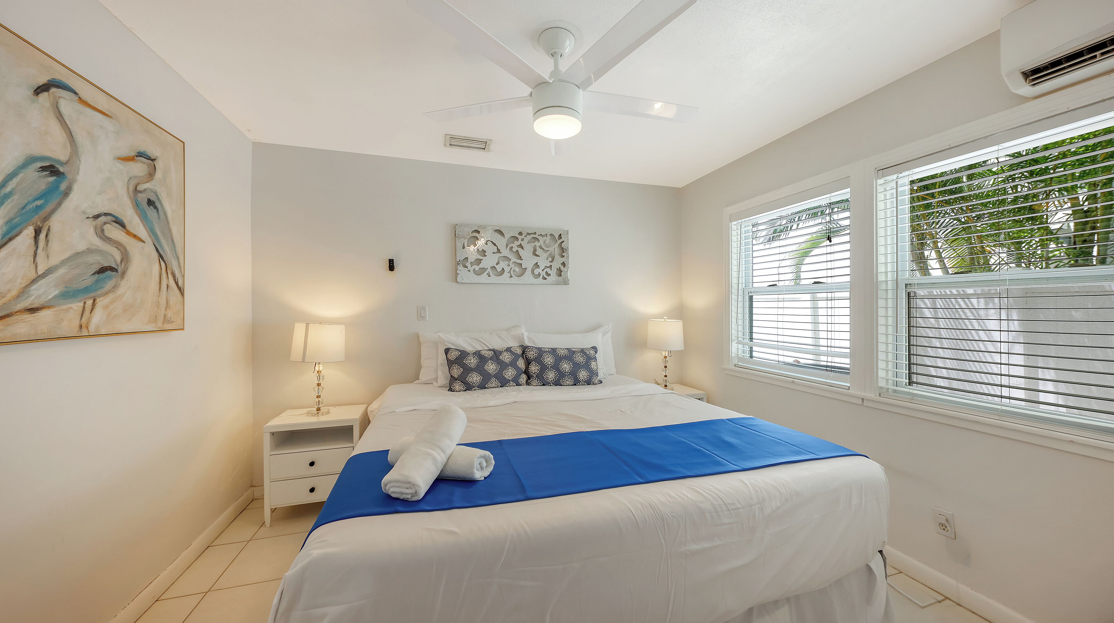 A bedroom with a bed, nightstands, a ceiling fan, a window with blinds, and a wall art piece.