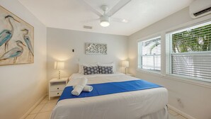 Een slaapkamer met een bed, nachtkastjes, een plafondventilator, een raam met jaloezieën en een kunstwerk aan de muur.
