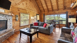 A wooden cabin interior with a stone fireplace, a gray sofa with red cushions, a wooden dining table, and a view through large windows. - Sleeps 30 | 5 Cabins + Hot Tubs + Firepit + Views | Mtn Views + Smokies Access (Townsend)