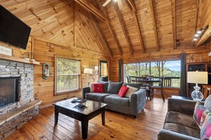 A wooden cabin interior with a stone fireplace, a gray sofa with red cushions, a wooden dining table, and a view through large windows. - Sleeps 30 | 5 Cabins + Hot Tubs + Firepit + Views | Mtn Views + Smokies Access (Townsend)
