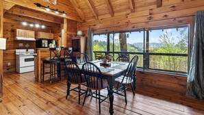 A wooden cabin with a dining area, kitchen, and large windows overlooking a forest. - Sleeps 30 | 5 Cabins + Hot Tubs + Firepit + Views | Mtn Views + Smokies Access (Townsend)