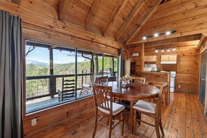 Una cabaña de madera con una mesa de comedor y sillas, una zona de cocina y ventanas amplias que ofrecen vistas al exterior.