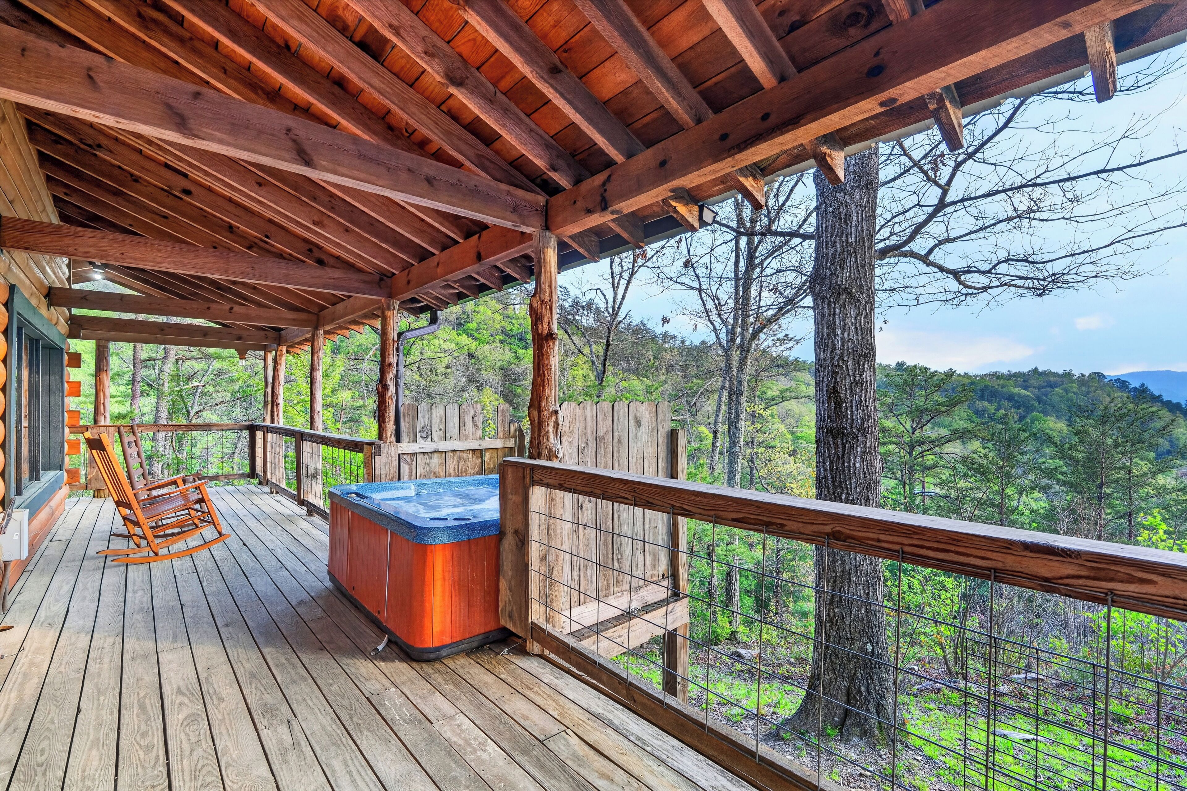 Een houten terras met een jacuzzi, schommelstoelen en uitzicht op een bebost landschap.