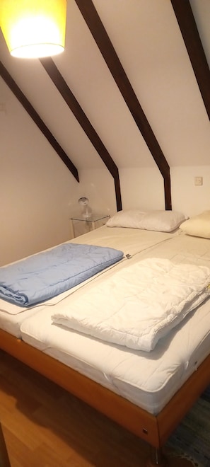 Ein Einzelbett mit weißer Bettwäsche, einer blauen Decke und einem Holz-Kopfstück in einem Raum mit schrägen Decken und einer sichtbaren Lampe.