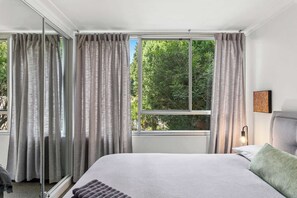 A bedroom with a large window, a bed, and a bedside table with a lamp. - Cosy Darling Point apartment (Darling Point)