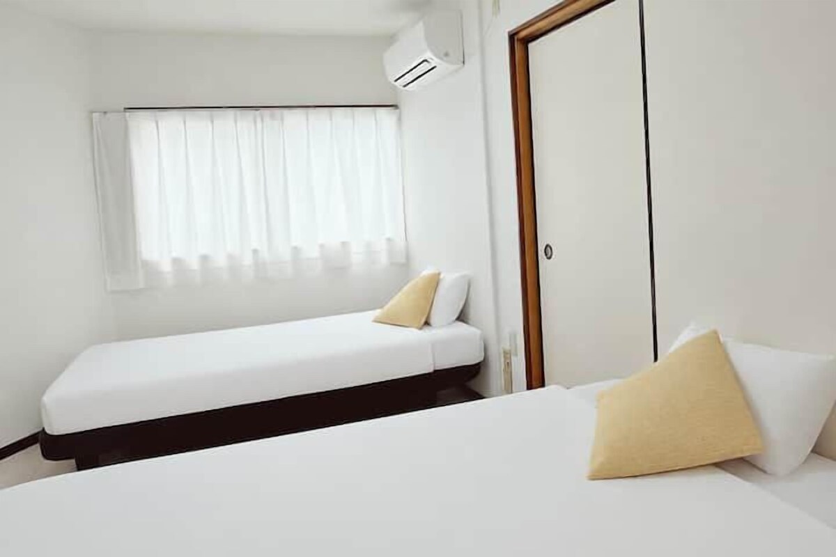 Habitació d'hotel amb dos llits, una finestra amb cortines, una porta i un aparell d'aire condicionat.