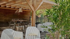 Un’area salotto esterna con sedie bianche e un tavolo di legno, sotto un pergolato di legno con luci a sospensione.