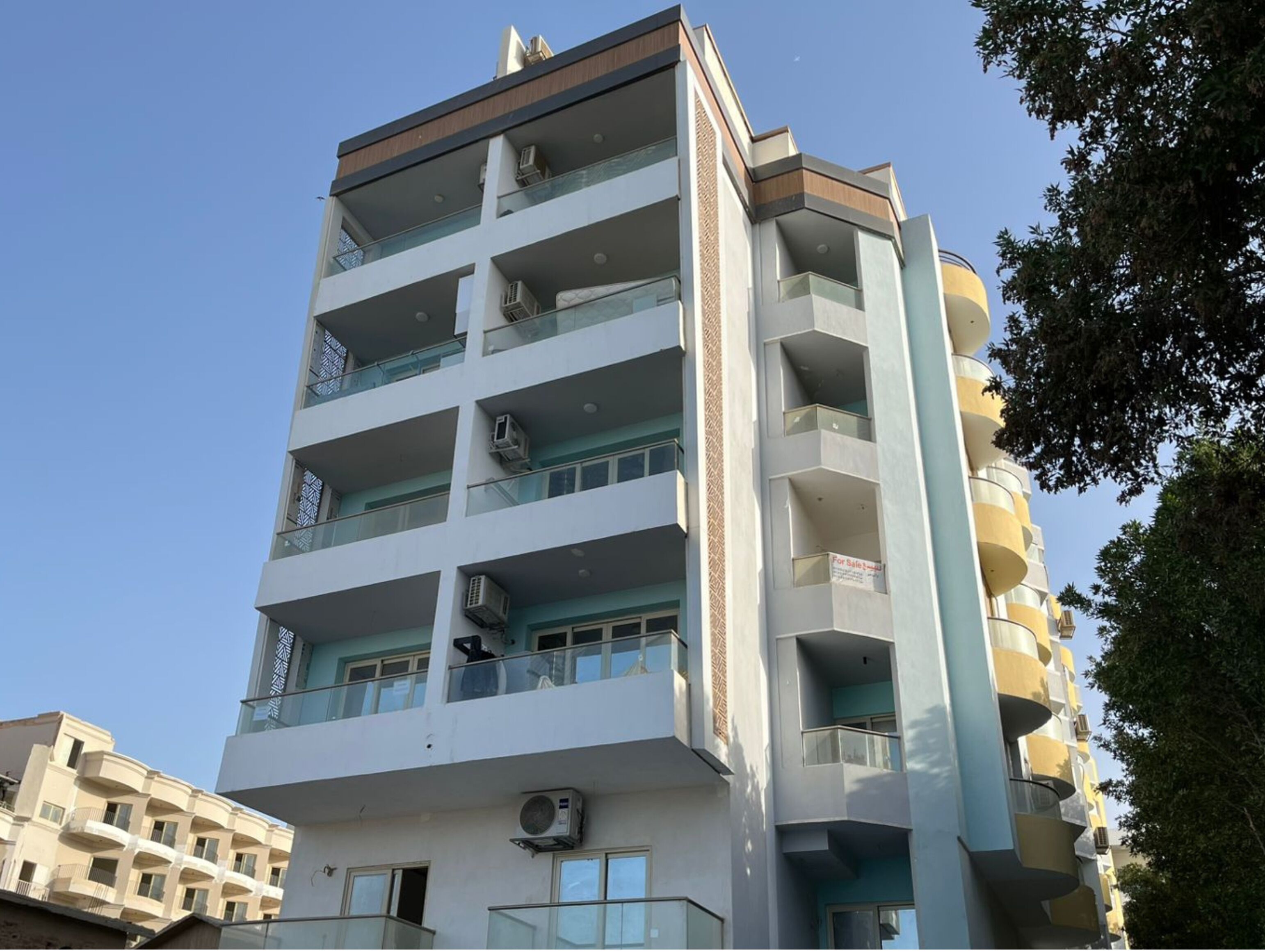 Un edificio residencial de varios pisos con balcones, unidades de aire acondicionado y un cartel de 'For Sale'.