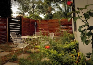 Eine Terrasse mit zwei weißen Metallstühlen und einem Tisch, umgeben von Grünpflanzen und einem Holzzaun.
