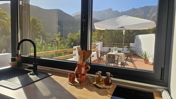 Uma cozinha com janela, utensílios de madeira e uma mesa com guarda-sol branco.