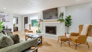 A living room with a fireplace, a sofa, a coffee table, and a TV mounted on the wall. - GREEN LANTERN: Game Room, Furnished Deck, Modern (Lexington)