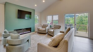 A spacious living area with a flat-screen TV, comfortable seating, a fireplace, and a view of the outdoors. - Margaritaville Access! Luxe Ozark Oasis w/ Hot Tub (Osage Beach)