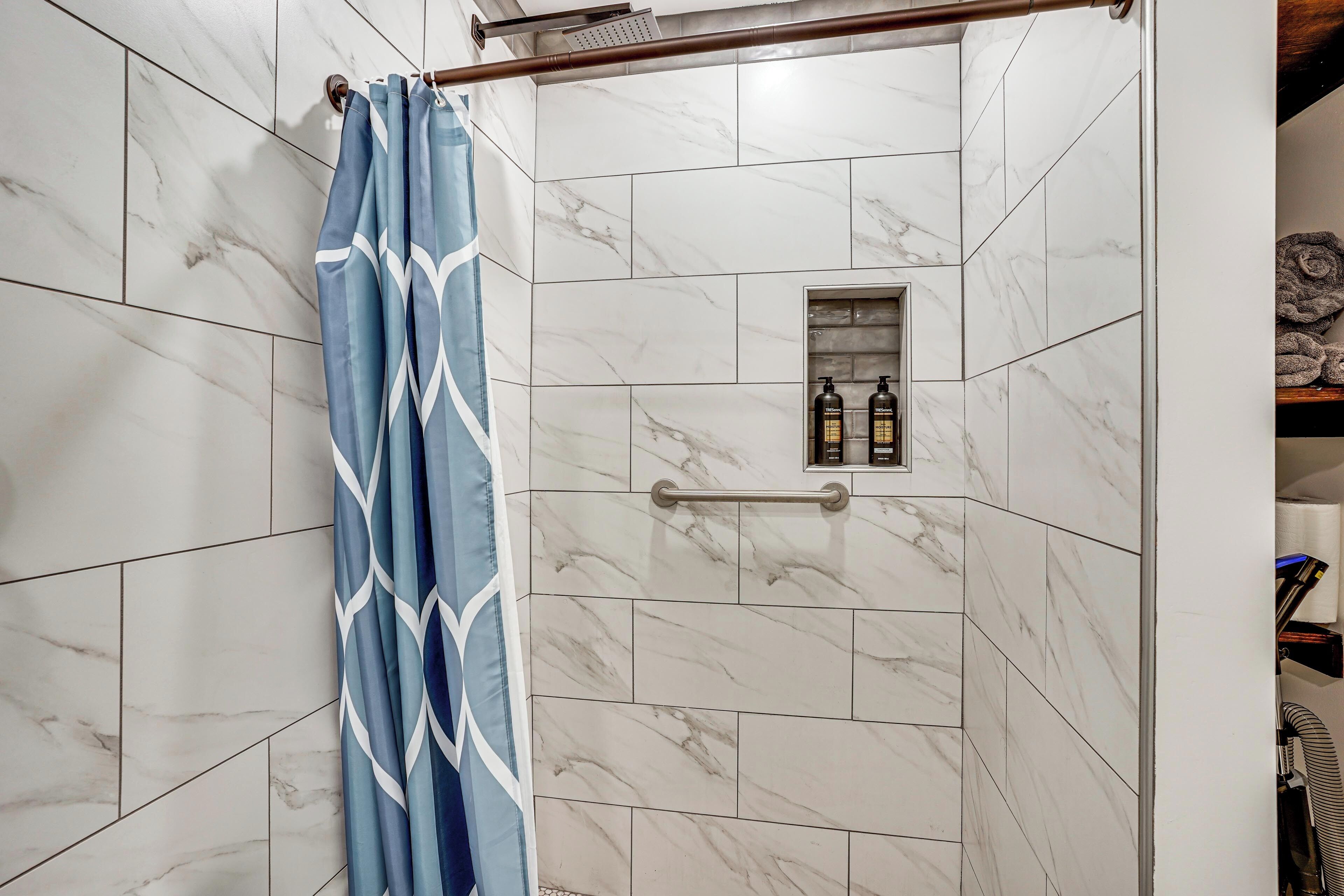 A shower area with a blue patterned shower curtain, a small shelf with bottles, and a handrail.