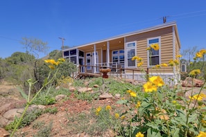 Une maison à deux étages avec un porche, entourée d’un jardin agrémenté de fleurs jaunes et de rochers.