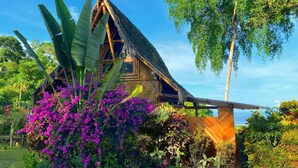 A thatched-roof house with a wooden deck, surrounded by lush greenery and flowering plants.