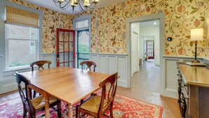 Un coin repas avec une table et des chaises en bois, un tapis rouge à motifs et un mur recouvert d’un papier peint à motifs de fruits et de fleurs.